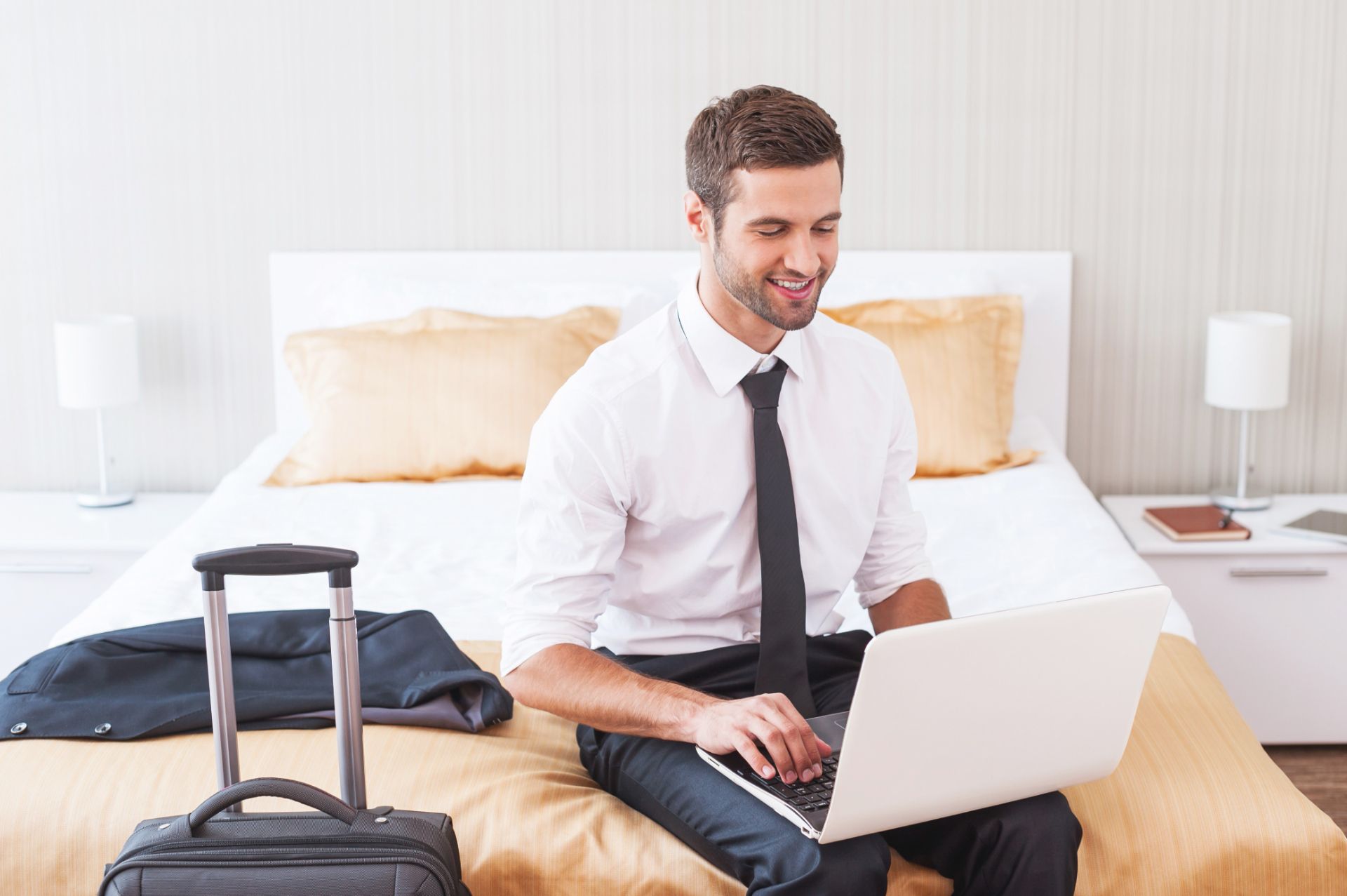 Homem sorrindo sentado na cama com notebook no colo e mala ao lado